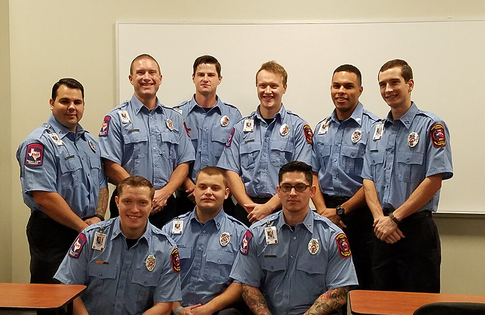 Nine firefighters in uniform are posed in two rows inside a room with a whiteboard. They are smiling, with the front row seated and the back row standing. The uniforms are blue with various patches and badges.
