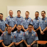 Nine firefighters in uniform are posed in two rows inside a room with a whiteboard. They are smiling, with the front row seated and the back row standing. The uniforms are blue with various patches and badges.
