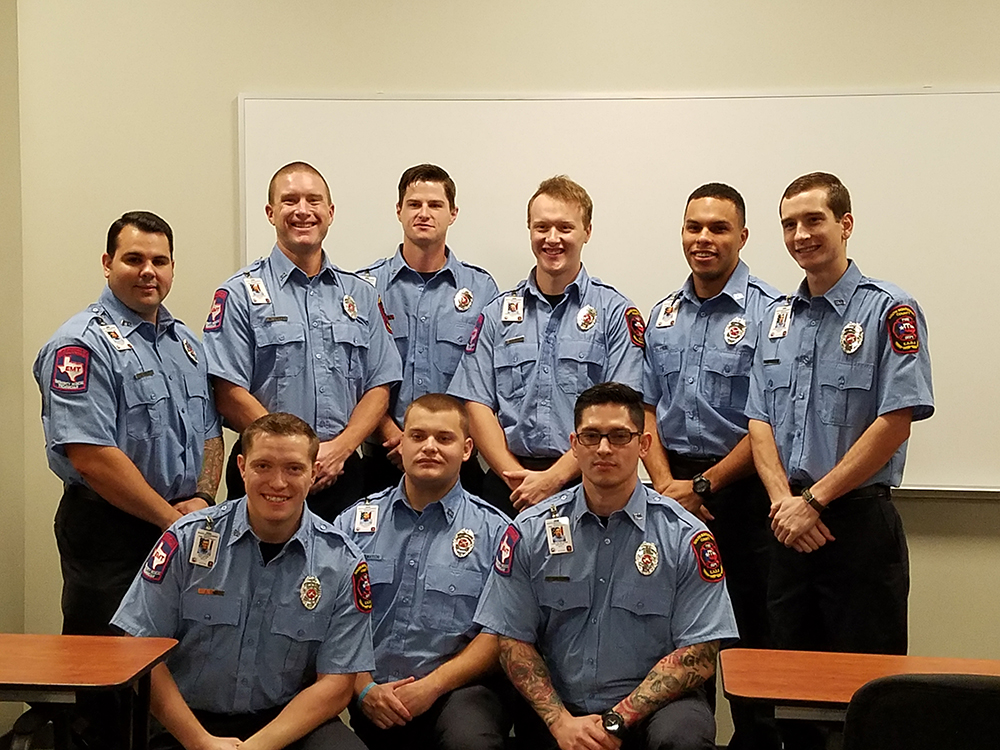 Nine firefighters in uniform are posed in two rows inside a room with a whiteboard. They are smiling, with the front row seated and the back row standing. The uniforms are blue with various patches and badges.