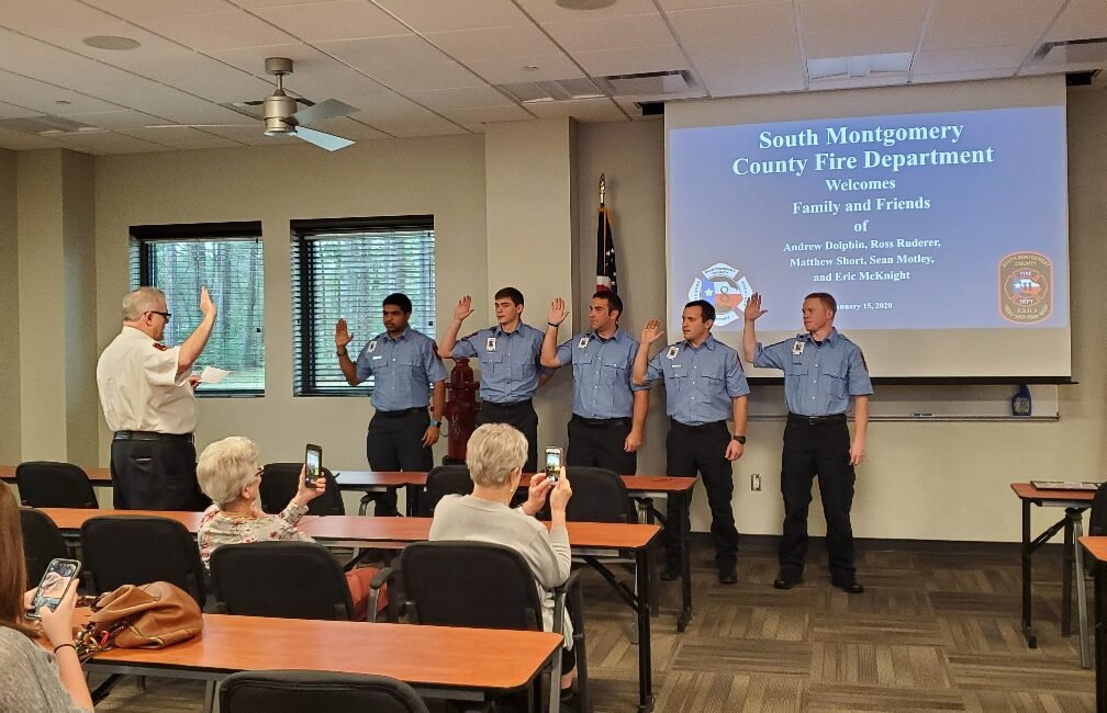 A group of five uniformed firefighters stands in front of an audience raising their right hands for an oath. A person to the left administers the oath. The screen displays information about the South Montgomery County Fire Department.