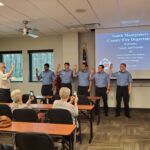 A group of five uniformed firefighters stands in front of an audience raising their right hands for an oath. A person to the left administers the oath. The screen displays information about the South Montgomery County Fire Department.