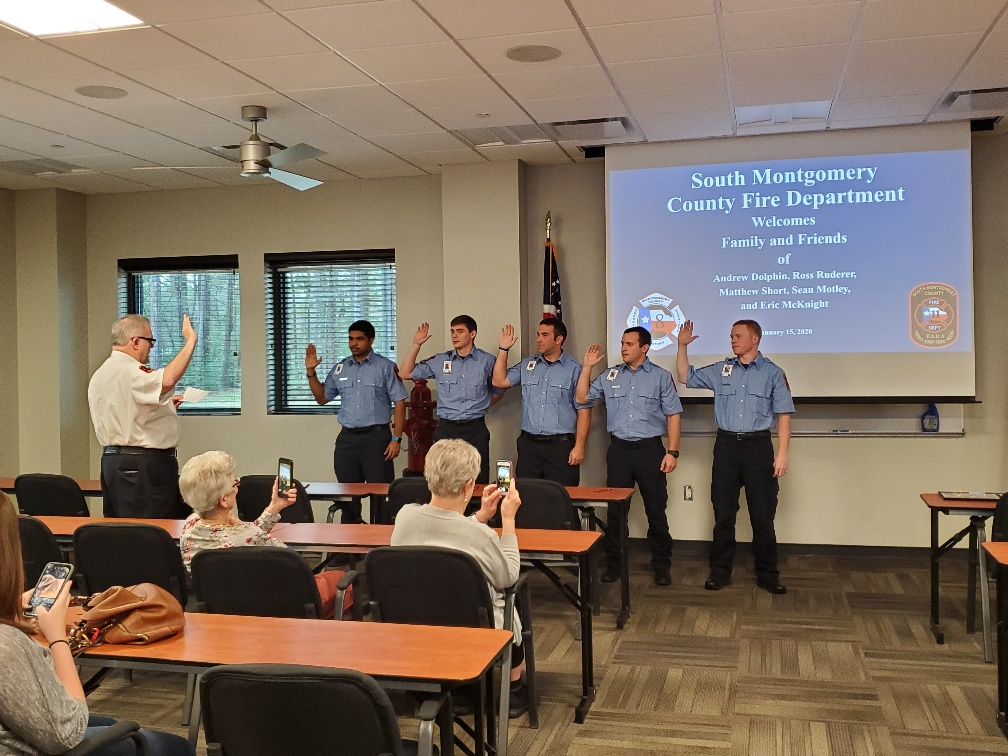 A group of five uniformed firefighters stands in front of an audience raising their right hands for an oath. A person to the left administers the oath. The screen displays information about the South Montgomery County Fire Department.