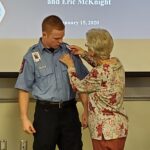 An elderly woman pins a badge on a young person dressed in a blue uniform during a ceremony. A presentation slide is visible in the background with names and a date, January 15, 2020.