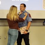 A person in a brown shirt pins a badge on a uniformed person holding a certificate. They are both smiling on a stage, with names and a logo projected on the screen behind them.