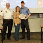 Three people stand indoors. A young man in a fire department uniform holds a certificate and smiles. A woman stands beside him, smiling with her arm around him. An older man in a uniform stands on the other side. A presentation is in the background.