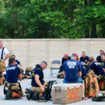 A group of firefighters in uniform rests outdoors, sitting on coolers and folding chairs. They are gathered in an open area with trees and a fence in the background. Their gear and equipment are scattered around.