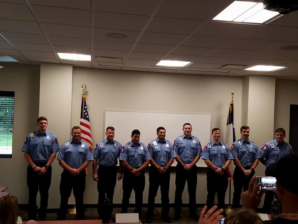 Nine people in uniform stand in a row in a room with an American flag and another flag. They are smiling, and some people are in the foreground taking photos. There is a whiteboard behind them on the wall.