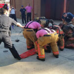 Group of firefighters in gear and a police officer assist in a rescue operation, using a stretcher outdoors. Fire truck visible in the background, with additional personnel nearby.