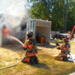 Three firefighters in gear are extinguishing a fire inside a shipping container outdoors. Smoke billows out, and flames are visible. Nearby are ladders, trees, and equipment. Two firefighters are kneeling, and one is standing with a hose.
