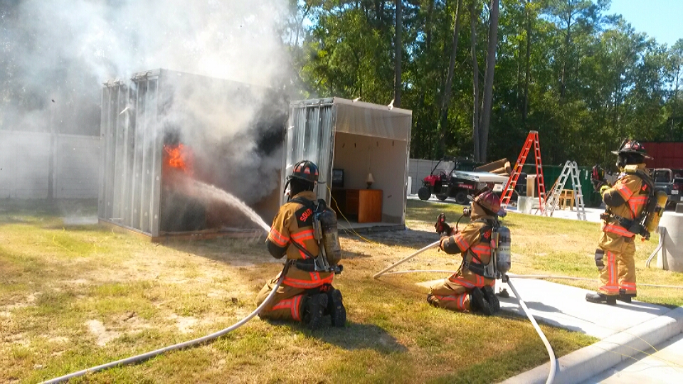 Three firefighters in gear are extinguishing a fire inside a shipping container outdoors. Smoke billows out, and flames are visible. Nearby are ladders, trees, and equipment. Two firefighters are kneeling, and one is standing with a hose.