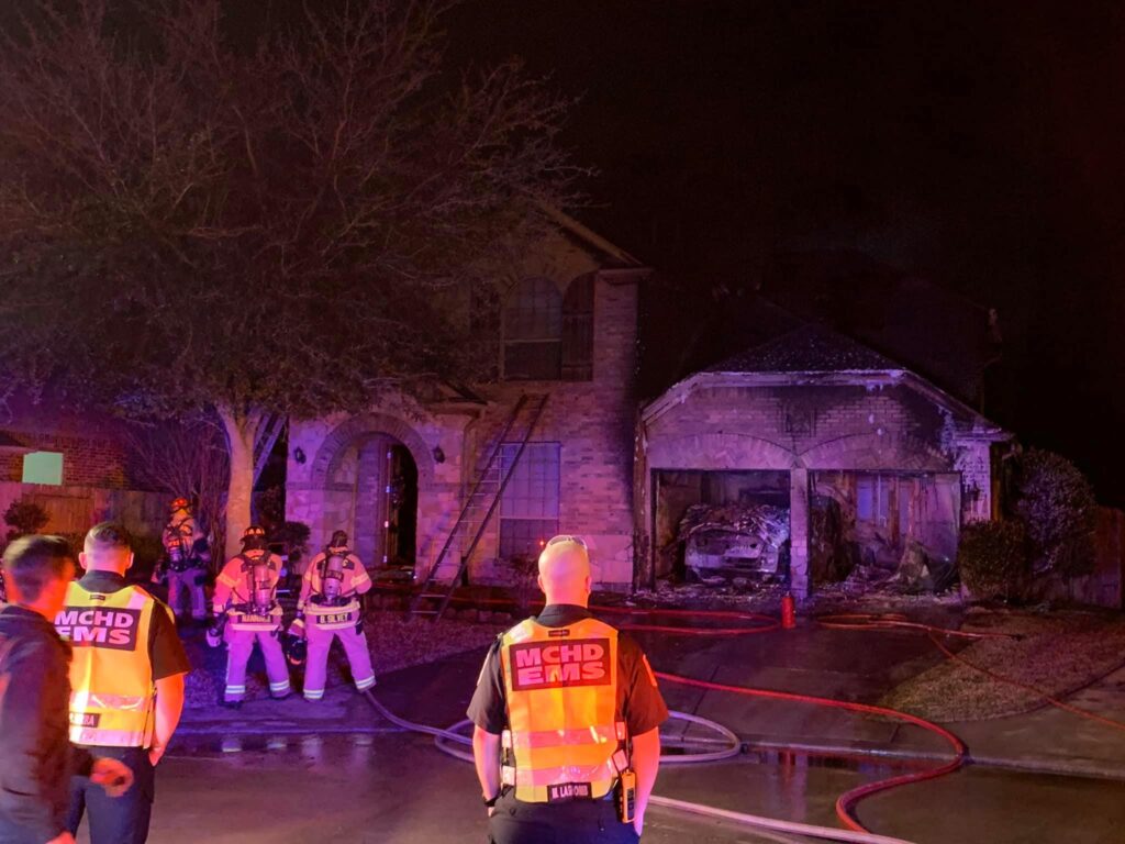 Firefighters and emergency personnel respond to a house fire at night. The brick house shows visible damage, with smoke and charred areas at the garage. Several hoses lie across the driveway, and emergency people wear reflective vests.
