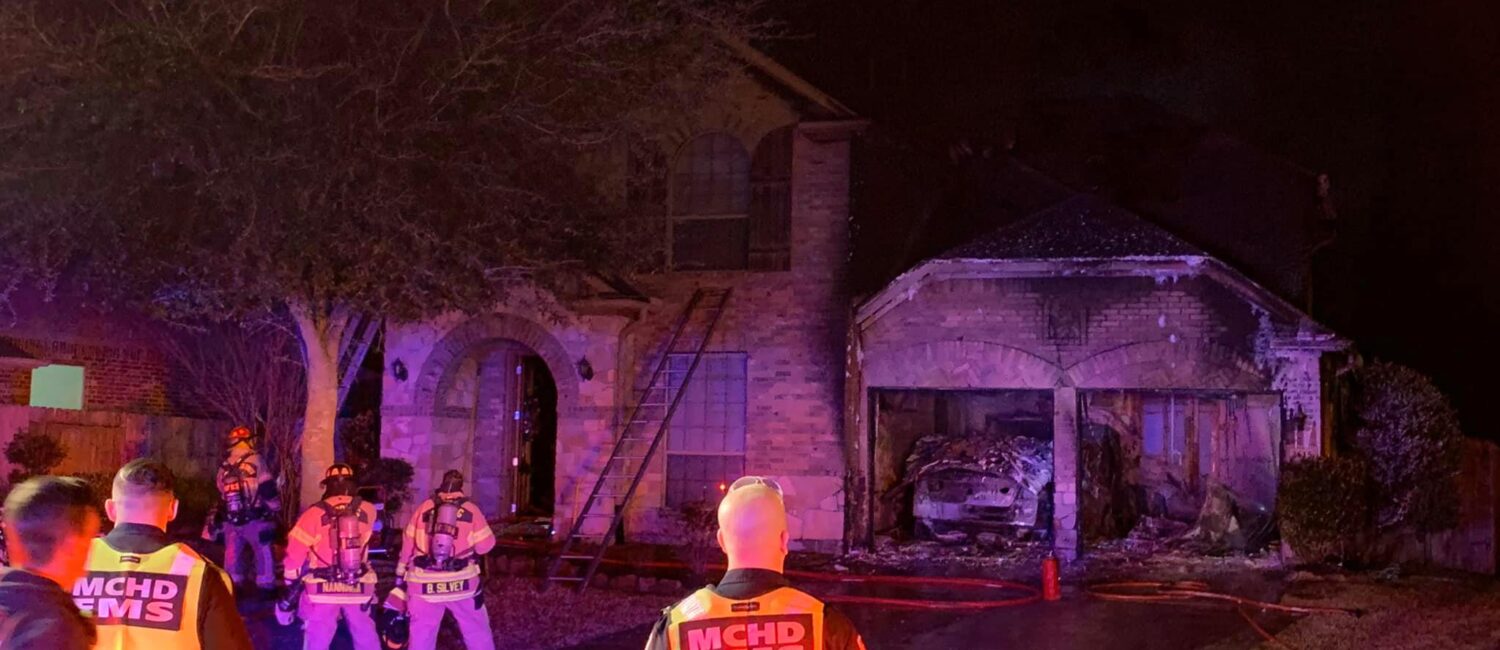 Firefighters and emergency personnel respond to a house fire at night. The brick house shows visible damage, with smoke and charred areas at the garage. Several hoses lie across the driveway, and emergency people wear reflective vests.