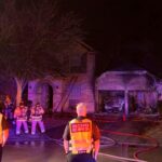Firefighters and emergency personnel respond to a house fire at night. The brick house shows visible damage, with smoke and charred areas at the garage. Several hoses lie across the driveway, and emergency people wear reflective vests.