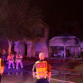Firefighters and emergency personnel respond to a house fire at night. The brick house shows visible damage, with smoke and charred areas at the garage. Several hoses lie across the driveway, and emergency people wear reflective vests.