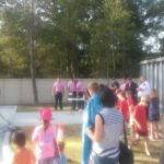 A group of people, including children, gather outdoors watching firefighters in gear demonstrate by a car. Trees and a container are in the background. A few men in pink shirts stand near a table. The scene is sunny and informal.