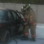 Two firefighters in gear work on opening a black cars door using a hydraulic rescue tool. They are in a paved area near a concrete wall on a sunny day.