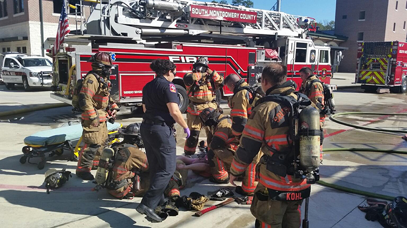 Firefighters, in full gear, gather around a firefighter being assisted on the ground at a fire station. A fire truck with a ladder is parked nearby. The scene appears to be a training or rescue situation.