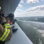 Firefighters in protective gear spray water from a hose over a high-rise balcony, aiming at an unseen target. The scene overlooks trees, buildings, and a partly cloudy sky in the distance.