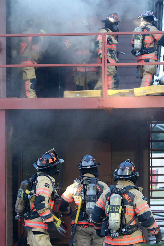 Firefighters in full gear, including helmets and oxygen tanks, are at the scene of a building fire. Some are on a balcony, while others are on the ground with hoses, surrounded by smoke, engaged in firefighting efforts.