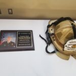 A retirement plaque and a beige firefighter helmet with a headlamp lay on a white table. The plaque features a photo of a firefighter in uniform and text marking the retirement of Deputy Chief Tommy Erickson, dated June 24, 2000 - June 27, 2020.
