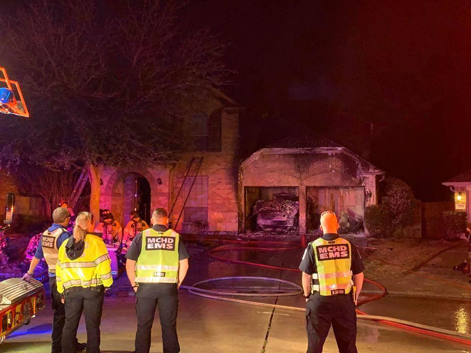 Emergency responders in reflective gear stand in front of a two-story house with a partially destroyed garage at night. Fire hoses lie on the wet driveway, and lights illuminate the scene.