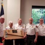 A group of five people in uniform pose indoors, holding a large wooden plaque with a golden baton and fire department insignia. An American flag is partially visible in the background beside a window showing greenery.