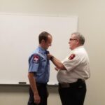 Two people in uniform are in front of a whiteboard. One is wearing a blue shirt with patches and is receiving an insignia or badge from the other person in a white shirt. The setting appears to be a formal or official ceremony.