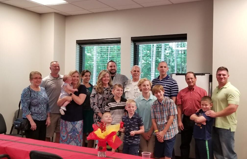 A group of people, including men, women, and children, are smiling and posing for a photo in a room with windows. A decorated table is in the foreground with a centerpiece.