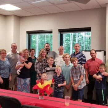 A group of people, including men, women, and children, are smiling and posing for a photo in a room with windows. A decorated table is in the foreground with a centerpiece.