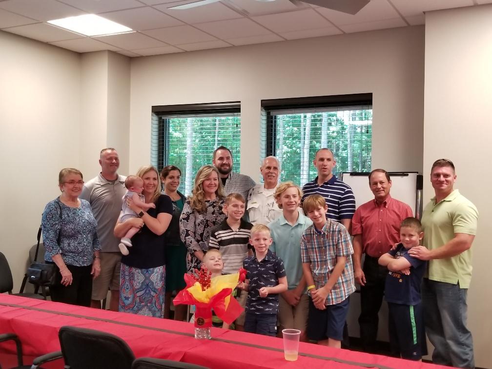 A group of people, including men, women, and children, are smiling and posing for a photo in a room with windows. A decorated table is in the foreground with a centerpiece.