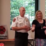 A man in a fire department uniform stands next to a woman holding a baby. They are indoors, near a window with trees outside. A sign with crossed axes is on a table behind them. The woman gestures with one hand.
