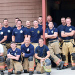 A group of firefighters poses for a photo. They are wearing matching navy blue T-shirts, with some in tan and reflective-striped firefighting pants. They stand in front of a brown building, smiling at the camera.