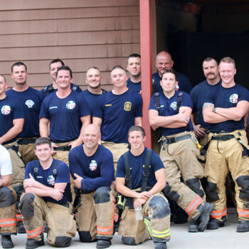A group of firefighters poses for a photo. They are wearing matching navy blue T-shirts, with some in tan and reflective-striped firefighting pants. They stand in front of a brown building, smiling at the camera.