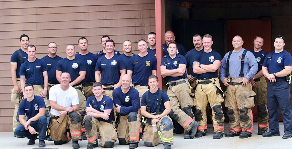 A group of firefighters poses for a photo. They are wearing matching navy blue T-shirts, with some in tan and reflective-striped firefighting pants. They stand in front of a brown building, smiling at the camera.