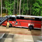 A red fire truck with NEEDHAM written on its side is parked near a building with green trees in the background. A firefighter is standing at the rear of the truck. The scene is sunny and the ground is wet.
