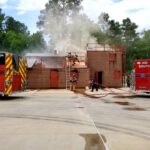 Firefighters conduct a training exercise at a smoke-filled structure. Two fire trucks are parked nearby. One firefighter holds a hose, while others work on the roof. The scene is set in a wooded area with a clear sky.