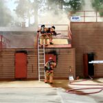 Firefighters in full gear participate in a training exercise. Two are on a structure with smoke, one ascending a ladder, and another standing nearby with hoses. A wall-mounted sign reads Dräger. A person in a shirt marked FIRE stands nearby.