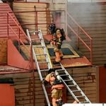 Two firefighters in gear climb a metal ladder leading to a buildings rooftop, surrounded by light smoke. The structure is brown with red accents. One firefighter is on the roof, and the other is partway up the ladder.