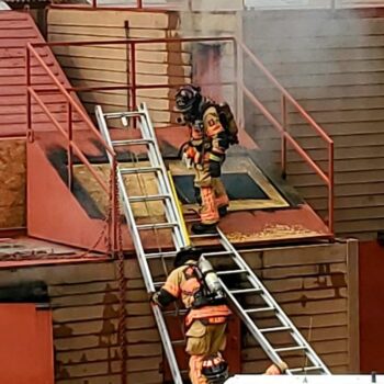 Two firefighters in gear climb a metal ladder leading to a buildings rooftop, surrounded by light smoke. The structure is brown with red accents. One firefighter is on the roof, and the other is partway up the ladder.