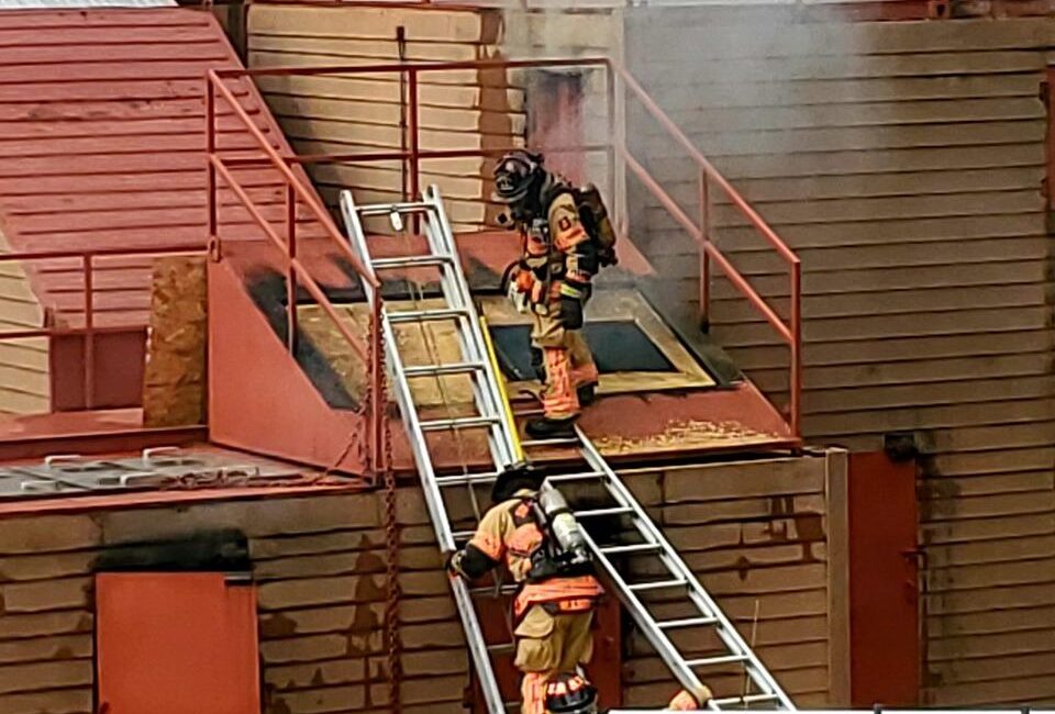 Two firefighters in gear climb a metal ladder leading to a buildings rooftop, surrounded by light smoke. The structure is brown with red accents. One firefighter is on the roof, and the other is partway up the ladder.