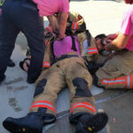 Firefighters perform a team-building exercise, assisting a colleague in turnout gear lying on his back. Two firefighters wear pink shirts and pants, one is securing straps on the ground, while another adjusts the equipment and helmet.