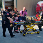 Emergency responders assist a person on a stretcher outside a fire station. People in uniforms and protective gear are gathered nearby. A red emergency vehicle is parked in the background.