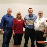Four people stand in a classroom. One person in a light blue shirt holds a certificate, smiling. Two people stand to the left, one in a red blouse and another in a blue shirt. A person in a white uniform stands to the right. A whiteboard is in the background.