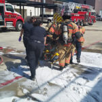 Firefighters and a medical professional assist a person on a stretcher outside a fire station. Foam covers the ground, and fire trucks are parked nearby.