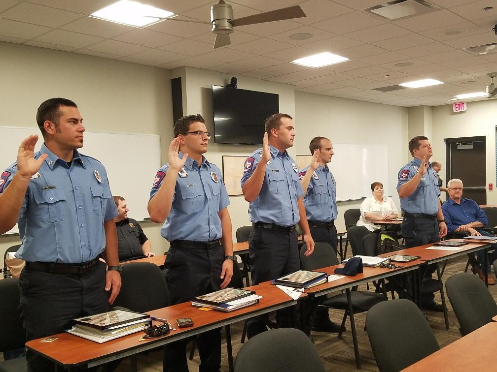 Five men in blue uniforms stand with their right hands raised in a classroom setting, taking an oath. Seated individuals watch, and tables hold binders and caps. Whiteboards and a screen are in the background.