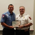 Two men in uniform stand together, smiling as they hold a Certificate of Appreciation. The man on the left wears a blue shirt, while the man on the right wears a white one. They are in front of a light-colored wall.