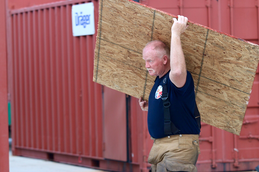 A person in a navy blue shirt and khaki pants carries a large wooden board on their shoulders. They are walking past red shipping containers.