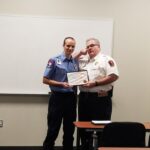 A man in a blue uniform stands beside a man in a white uniform, both holding a certificate. They are in a classroom setting with a whiteboard behind them. The man in white gestures with his hand near his forehead, as if saluting.