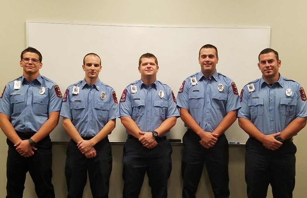 Five uniformed firefighters stand in a row in front of a whiteboard, wearing light blue shirts with badges and dark pants. They are posing for a group photo indoors.