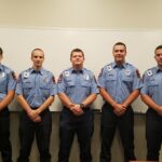 Five uniformed firefighters stand in a row in front of a whiteboard, wearing light blue shirts with badges and dark pants. They are posing for a group photo indoors.